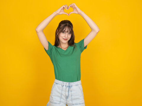 Pretty young woman making heart shape with hands over her head and smiling, standing against bright yellow background. Concept of love, health, happiness, kindness and supporting community.
