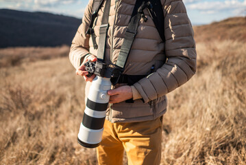 Close-up of an outdoor photographer stands among dry grasslands, using a long zoom lens to photograph nature on a clear day. © djoronimo