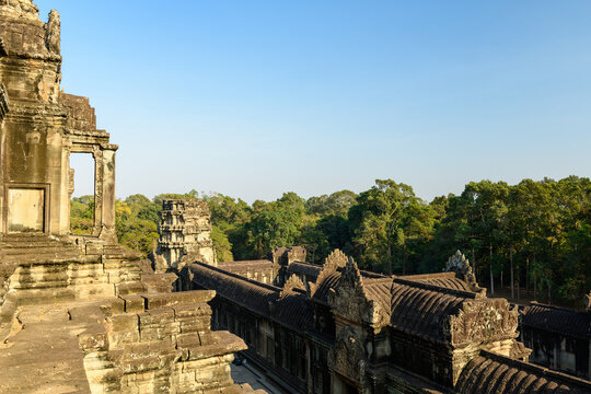 Warm afternoon light bathes the weathered sandstone steps and ornate rooflines at the summit of Angkor Vat, Cambodia. The ancient Khmer architecture stands above dense tropical forest under a clear