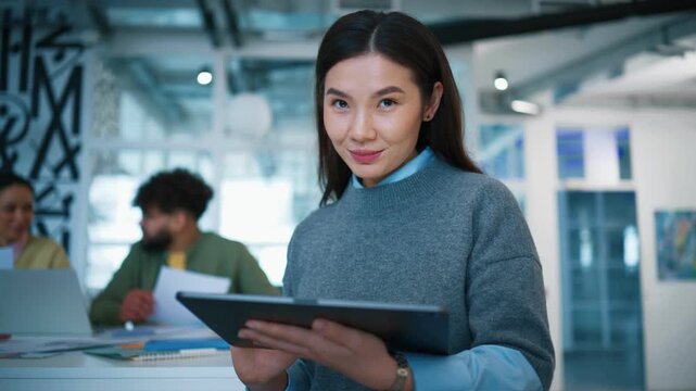 Cheerful Asian businesswoman holding tablet device. Smiling female looking at camera. Curly Caucasian male colleague reviewing papers. Team collaborating inside contemporary startup office.
