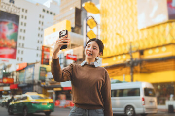 Happy Asian woman traveler taking selfie with smartphone at Yaowarat Road, young lady enjoying street food and neon lights at Bangkok Chinatown night market, concept of Thailand travel.