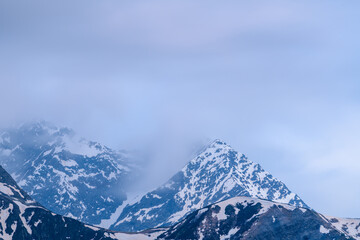 Mont Lachat's pointed summit emerges through drifting mist, with streaks of melting snow on rugged slopes. Soft blue light and overcast sky create a tranquil, ethereal alpine mood. © Florent