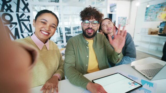 Group colleagues posing for selfie at desk. Female worker holding camera with one hand. Team members waving and smiling. Business people greeting viewer inside creative office space.