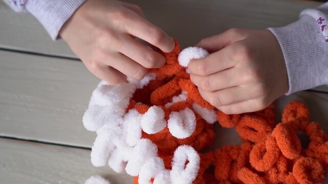 Little baby's fingers are knitted using white and orange plush yarn.. Children's hobby. The girl sits holding a woolen thread in her hands.