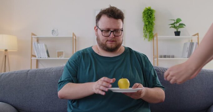 Fat overweight man with glasses sits on a sofa studying an apple placed on a plate in a modern livingroom. Clear healthy choice for daily life. Focus on nutrition, diet, choice and lasting health.