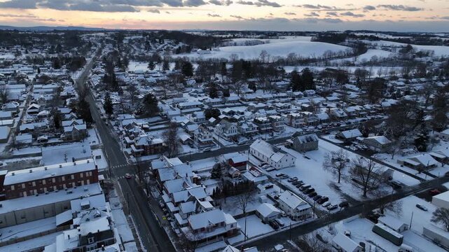 Aerial sunset view of Manheim, Pennsylvania covered in fresh snow, showing small American town with residential streets, rooftops and surrounding farmland under a winter sky. Wide shot.