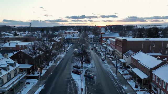 Aerial sunrise view of Manheim, Pennsylvania town center in winter with snow-covered rooftops, historic storefronts, quiet streets, parked cars and soft morning light over small American community.