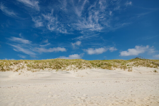 Scenic coastal landscape featuring a sandy beach with dry grass dunes and a wooden staircase leading to the sea under a bright blue sky with wispy cirrus clouds for travel and nature concepts