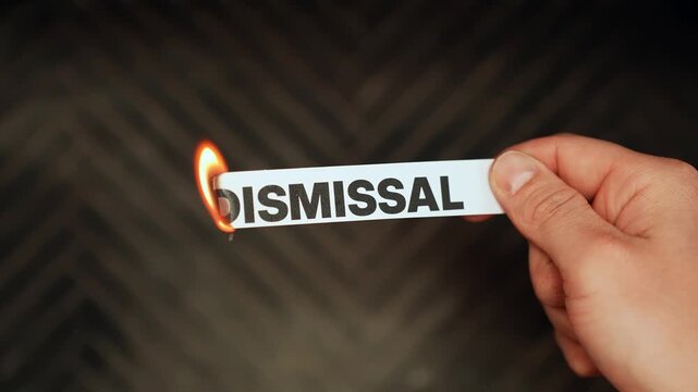 Close-up hand holds a paper strip labeled &ldquo;DISMISSAL&rdquo; as the edge burns with a flame on a dark blurred background&mdash;symbolic of rejection, stress, and workplace conflict, with copy space.