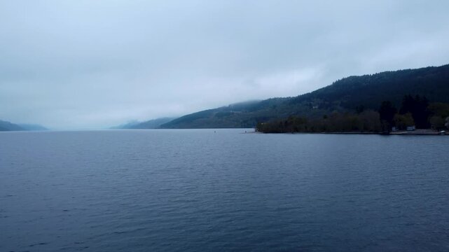 Tracking Drone Shot forward of the coast of Loch Ness in Scotland early in the morning against a cloudy sky. Only slightly above the water.