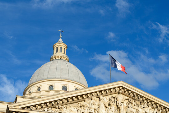 Close-up of the Pantheon dome in Paris with the French flag flying above ornate sculpted pediment details, set against a vivid blue sky with scattered clouds.