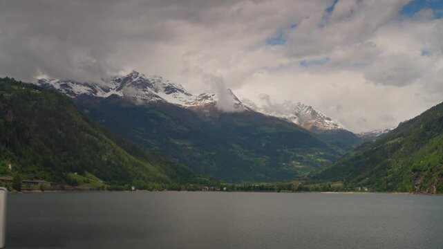 Panoramic view of Lake Poschiavo with snow capped Swiss Alps under cloudy sky in Val Poschiavo
