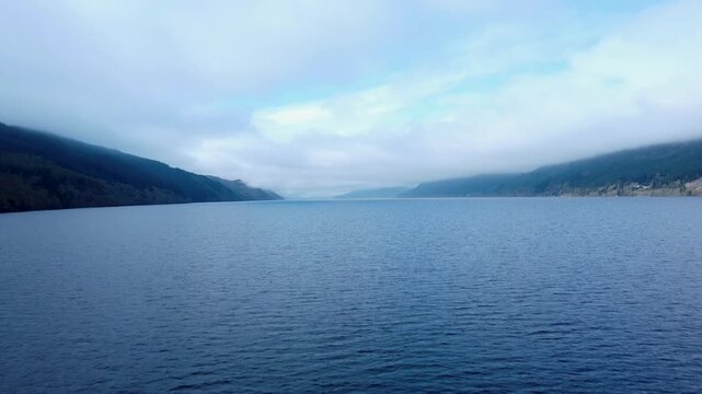 An Ascending Forward Track Drone Shot of Loch Ness Waters in Scotland forested mountains and hills rising in the distance meeting a cloudy summer sky.