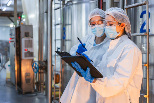 Female scientists wearing face masks and safety goggles working in high-standard processing facility, Quality control team using digital tablet and clipboard for production line audit in modern plant
