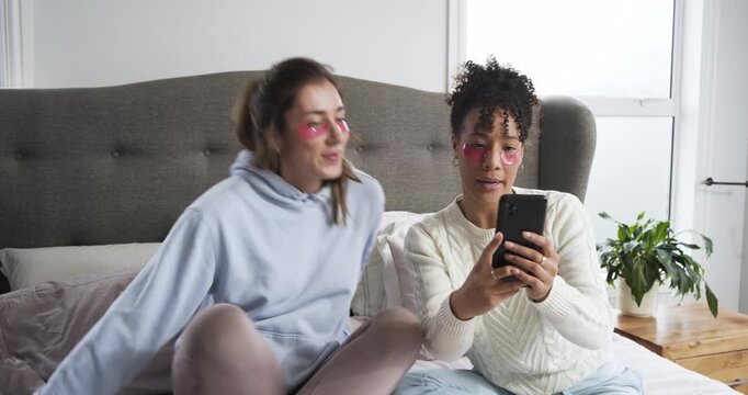 Diverse female friends applying eye patches on bed before posing and reviewing smartphone images