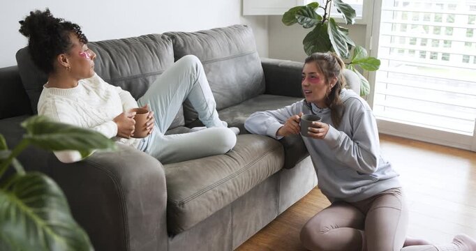After gel patch application, diverse female friends sipping mugs and chatting on sofa at home