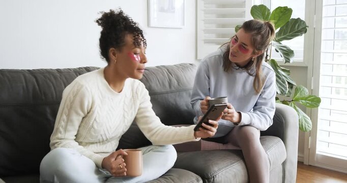 Diverse female friends laughing while swiping tablet on sofa after left friend raising it with mugs