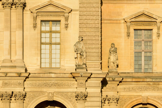 Two classical stone statues stand between tall pedimented windows on the ornate facade of the Louvre palace in Paris, France. Warm golden sunlight highlights the intricate carvings, textured masonry