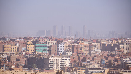 Obraz premium Densely built urban area with mid-rise residential buildings stretching across frame. Atmospheric haze softens distant skyline, where faint high-rise towers emerge in background. Giza and Cairo, Egypt