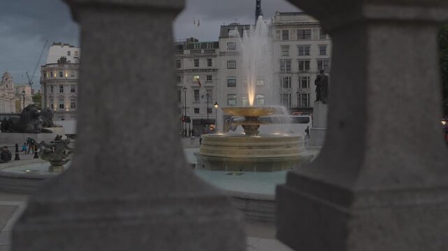 Artistic static shot of an illuminated fountain and the General Napier statue in Trafalgar Square, framed through stone railings at twilight.