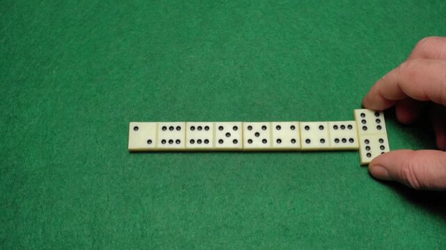 Hand placing domino piece onto existing row on green felt surface, showcasing the progression of a game with multiple dominoes aligned in sequence