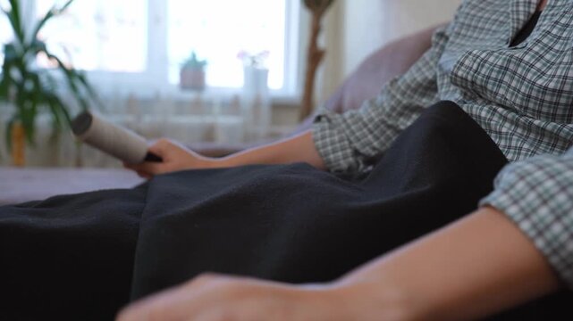 Woman's hand using a sticky lint roller to remove dust, hair, and lint from her black trousers while sitting on the sofa, demonstrating daily clothing care and household chores