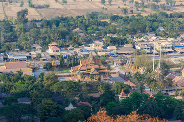 Fototapeta premium Elevated view of a traditional Buddhist temple with ornate golden roofs surrounded by lush trees, village houses, and dry agricultural fields near Phnom Sampov in Battambang province, Cambodia. The