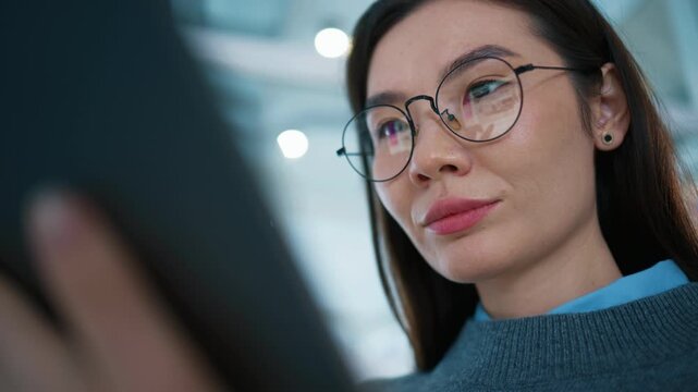 Close view of female employee using tablet with screen reflection visible in eyeglass lenses. Woman entering text on touch display while standing in bright corridor. Professional at work.