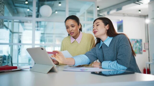 Team analysis session at office desk. Asian woman pointing at tablet screen and explaining data. Woman in yellow sweater holding document and listening carefully in modern workspace.