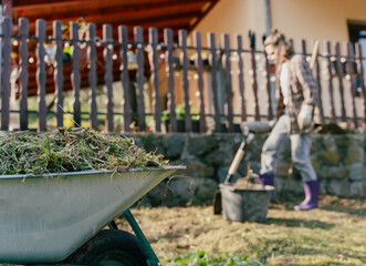 Wheelbarrow full of dry grass and weeds with blurred woman raking in the background of a country house.