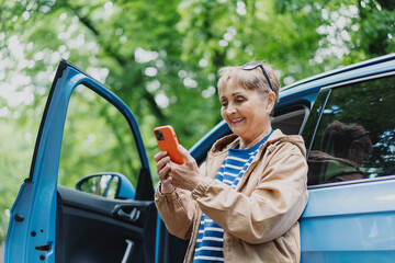 Happy senior woman driver standing by car and looking at smartphone on countryside road