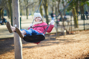 A young girl in a warm winter outfit having fun on a playground swing. Energetic outdoor childhood moment full of movement, joy, and active play.