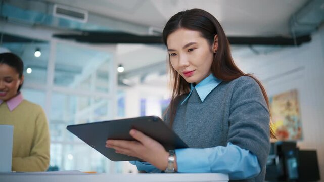 Focused office portrait at work desk. Asian woman holding tablet computer and reading digital information. Female professional checking online report while colleague smiling in background.