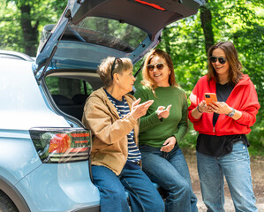 Happy senior mother and two adult daughters sitting in open car trunk and looking at smartphone