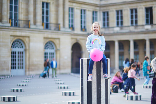 Smiling young girl balancing on one of the striped columns of the Buren installation at Palais-Royal in Paris, holding a bright pink balloon