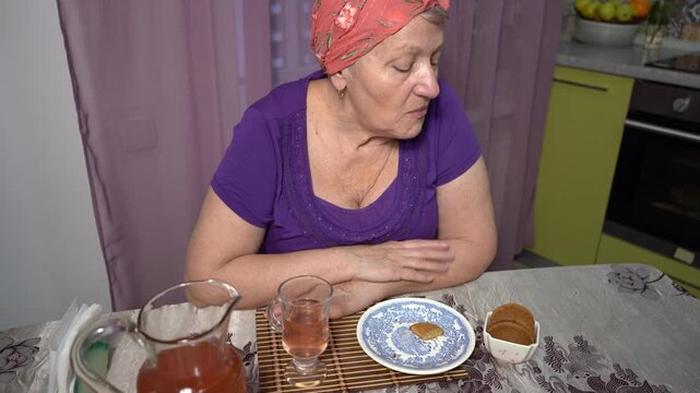 Elderly woman drinks compote from a glass at home. Pour a drink from a carafe. Eating cookies food