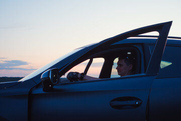 Naklejka premium Side view of male driver sitting in modern dark car with opened door holding steering wheel. Evening road trip concept with beautiful sunset sky background.
