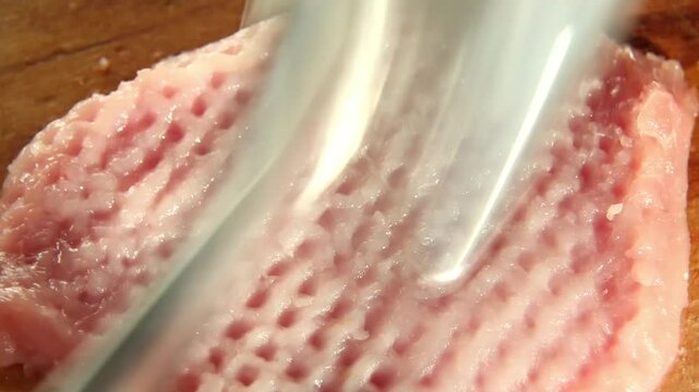 Overhead view of pork steak on a wooden board as metal tenderizer pounds repeatedly, pressing down then lifting, and dimpled texture spreads across the cutlet for quick, even cooking.