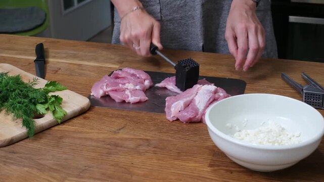 Hands using a black spiked meat hammer to tenderize raw pork slices on a metal board, kitchen preparation scene