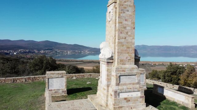 Close orbit drone shot of Doiran Memorial in northern Greece showing marble lion statue, engraved soldier names and wreath tribute with Lake Doiran and mountains in the distance.