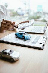 A woman reviews and signs a car insurance policy at her desk with a calculator, laptop, and...