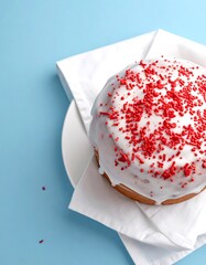 Round cake glazed white with red sprinkles, atop white napkin and plate on a blue backdrop