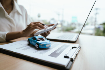 A woman reviews and signs a car insurance policy at her desk with a calculator, laptop, and miniature cars, representing auto coverage planning and financial protection. © Natthawadee