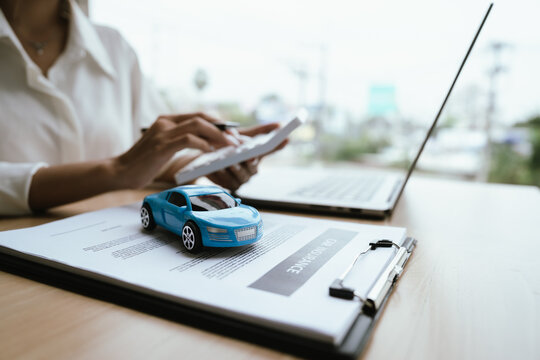 A woman reviews and signs a car insurance policy at her desk with a calculator, laptop, and miniature cars, representing auto coverage planning and financial protection.