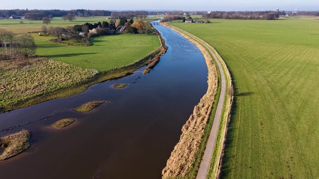 Aerial view of a scenic European river winding through green farmland, alongside a peaceful village featuring a historic brick church with a modern glass nave and houses with solar panels.