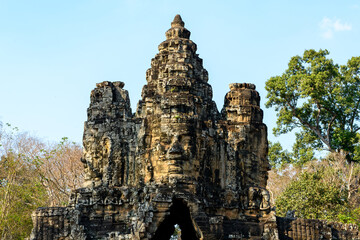 Naklejka premium Frontal view of a monumental stone gate crowned with serene carved faces at the entrance to Angkor Thom near Siem Reap, Cambodia. The ancient Khmer architecture rises above surrounding trees under
