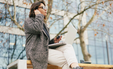 Mature business woman with laptop putting in wireless earbud for video call on a city bench