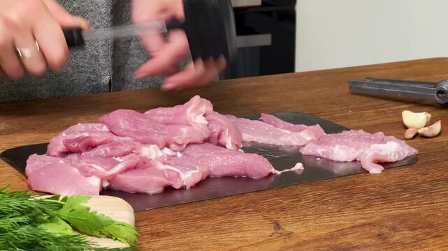 Hands using a black spiked meat hammer to tenderize raw pork slices on a metal board, kitchen preparation scene