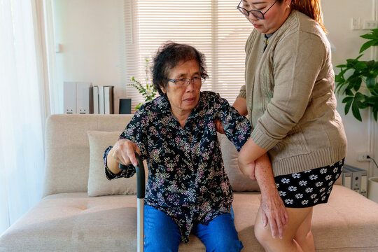 Young woman home caregiver providing empathy and support helping an elderly woman with walking stick getting up from sofa
