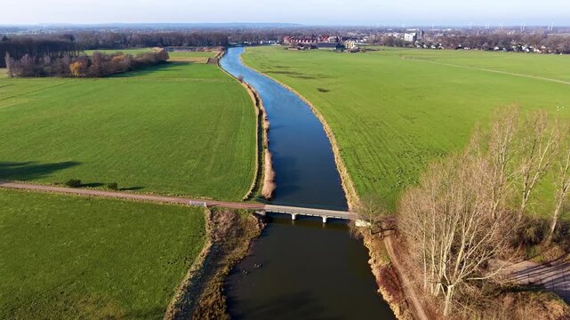 Aerial view of a rural landscape with a canal curving through green fields, two small road bridges crossing it, and a nearby house with outbuildings set among open farmland.